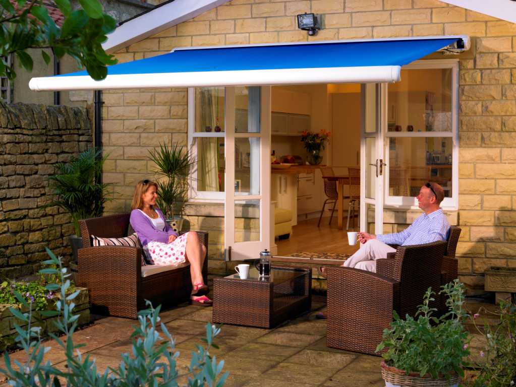 A couple dining in their garden on rattan furniture and covered by a blue awning